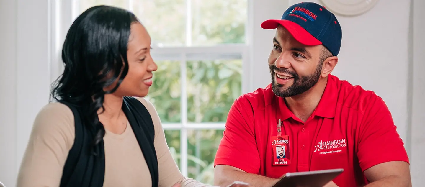 A Rainbow service professional sits beside a woman at a table, holding a tablet and explaining what is on it.