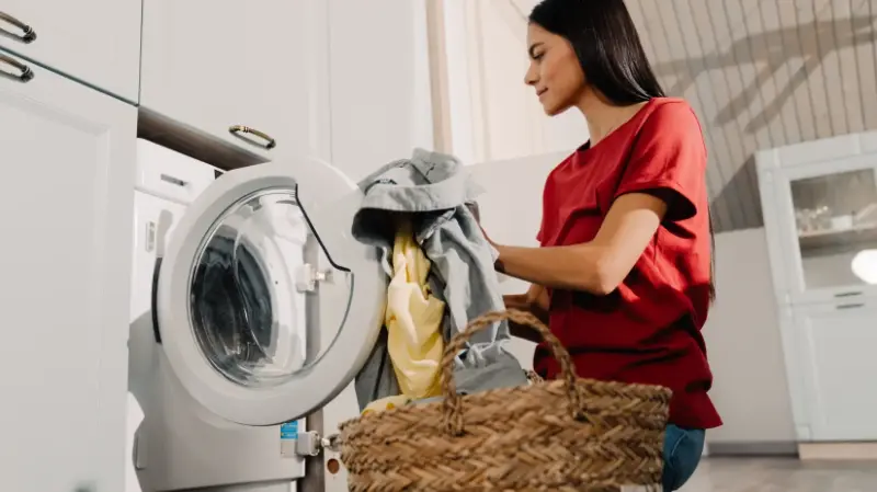 A woman sits in front of a washing machine filling it with clothes, alluding to the topic of how to remove smoke smell from clothes.
