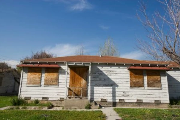 A single-story white house with boarded-up windows and front door, indicating damage or vacancy.