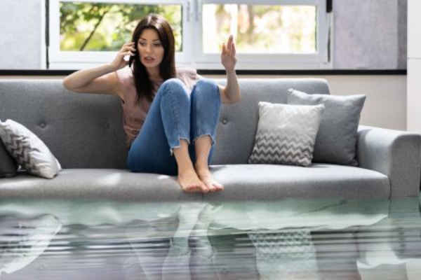 A woman sits on a gray couch with her legs pulled up, looking distressed as she talks on the phone. The room is flooded, with several inches of water covering the floor and reflecting the furniture. Sunlight filters through the windows behind her.