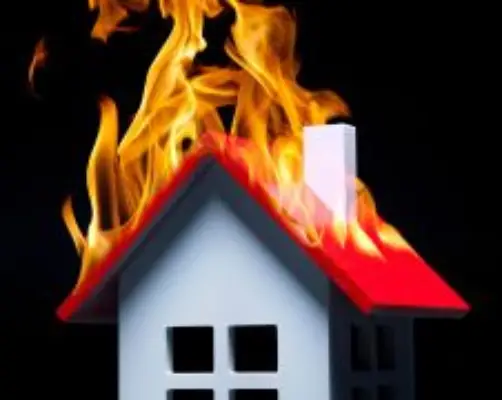 A small model house with a red roof is shown engulfed in bright orange flames against a dark black background, symbolizing a house fire.