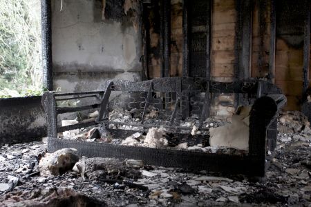 A severely fire-damaged living room with charred walls, a burned sofa frame, and debris scattered across the floor. The structure shows heavy smoke staining and destruction, highlighting the aftermath of a house fire.