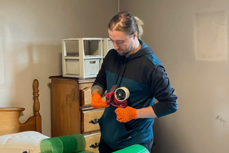 A restoration professional wearing orange gloves uses packing tape to secure protective wrap around household items in a bedroom. A wooden dresser and bed frame are visible as part of the content processing work.