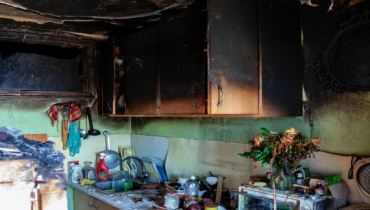 Kitchen damaged by a fire, and cabinets charred by fire, with black soot. A lot of items on the countertop.